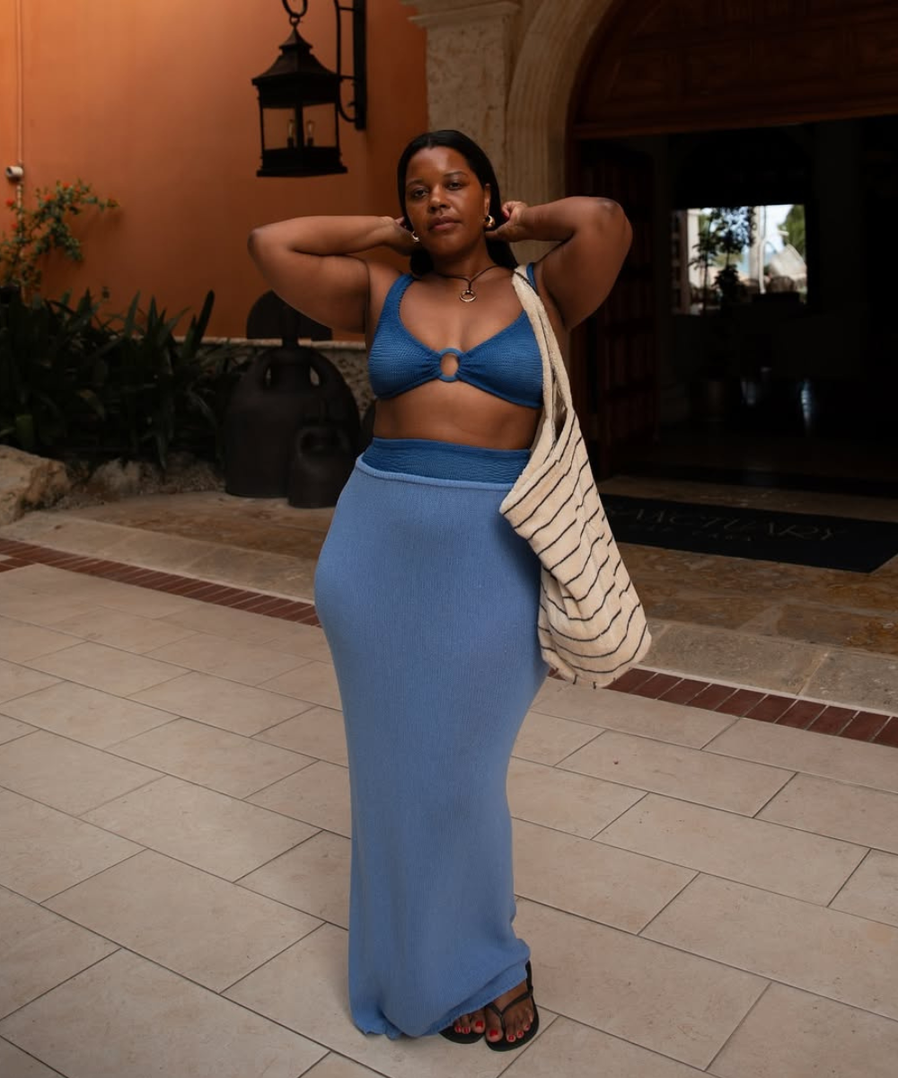 Modelo posa com top de crochê azul, saia longa ajustada e bolsa para praia de tecido cru com listras pretas.