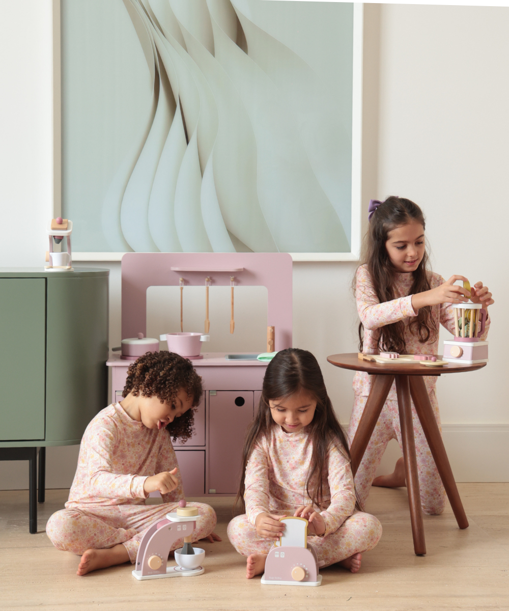 Três crianças brincam com brinquedos de madeira em tons pastel, destacando cozinha e eletrodomésticos em ambiente acolhedor.