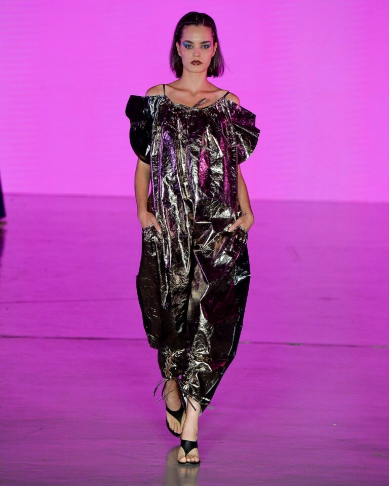 Modelo posa com vestido metalizado prateado de modelagem ampla, ombros à mostra e acabamento franzido no tornozelo, em clima