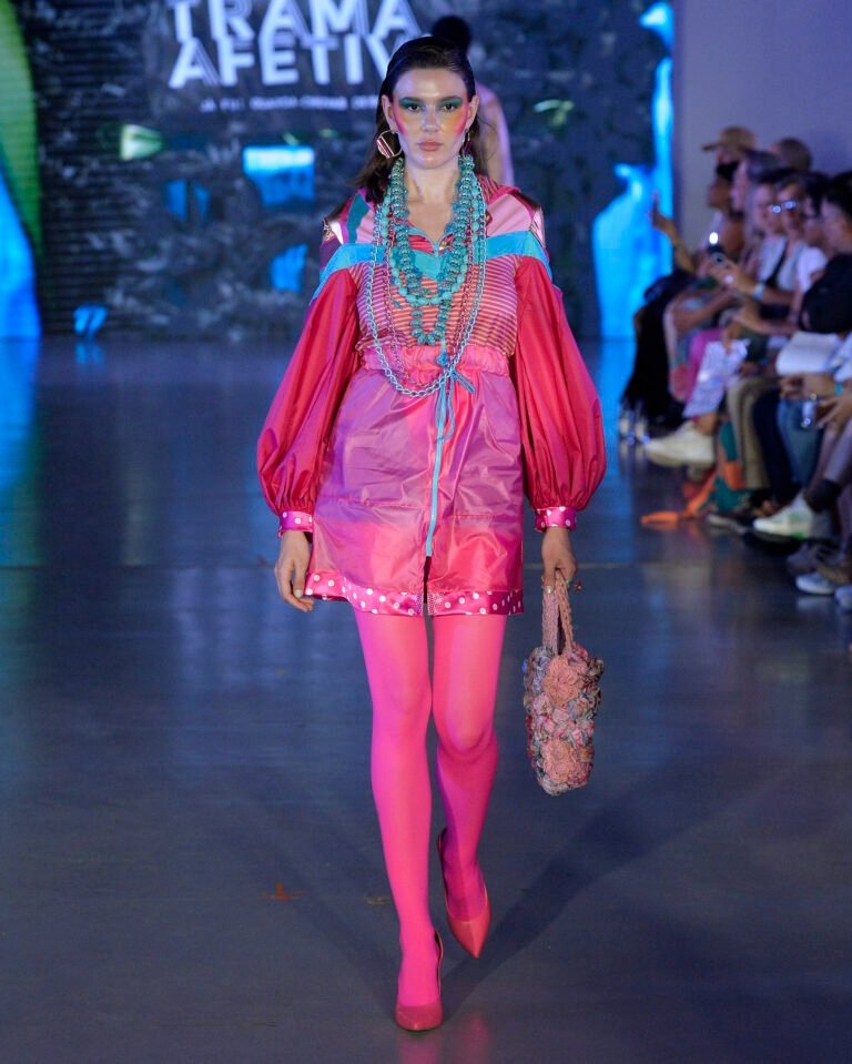 Modelo posa com vestido-capa rosa vibrante de tecido leve, mangas bufantes, meias e scarpins pink, colares turquesa e bolsa