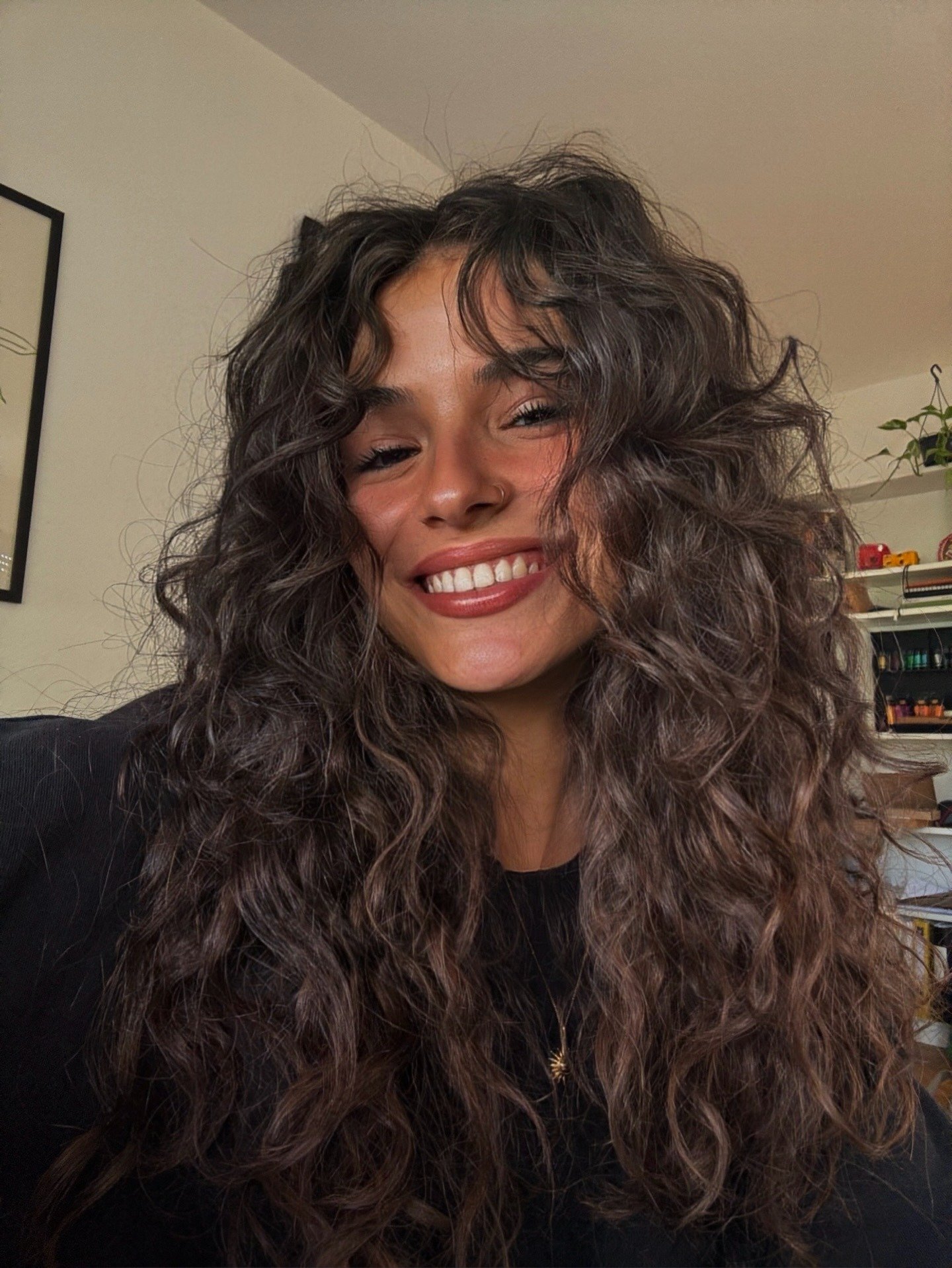 Mulher sorri com cabelo ondulado volumoso em tons castanhos, blusa preta e colar delicado, expressando alegria