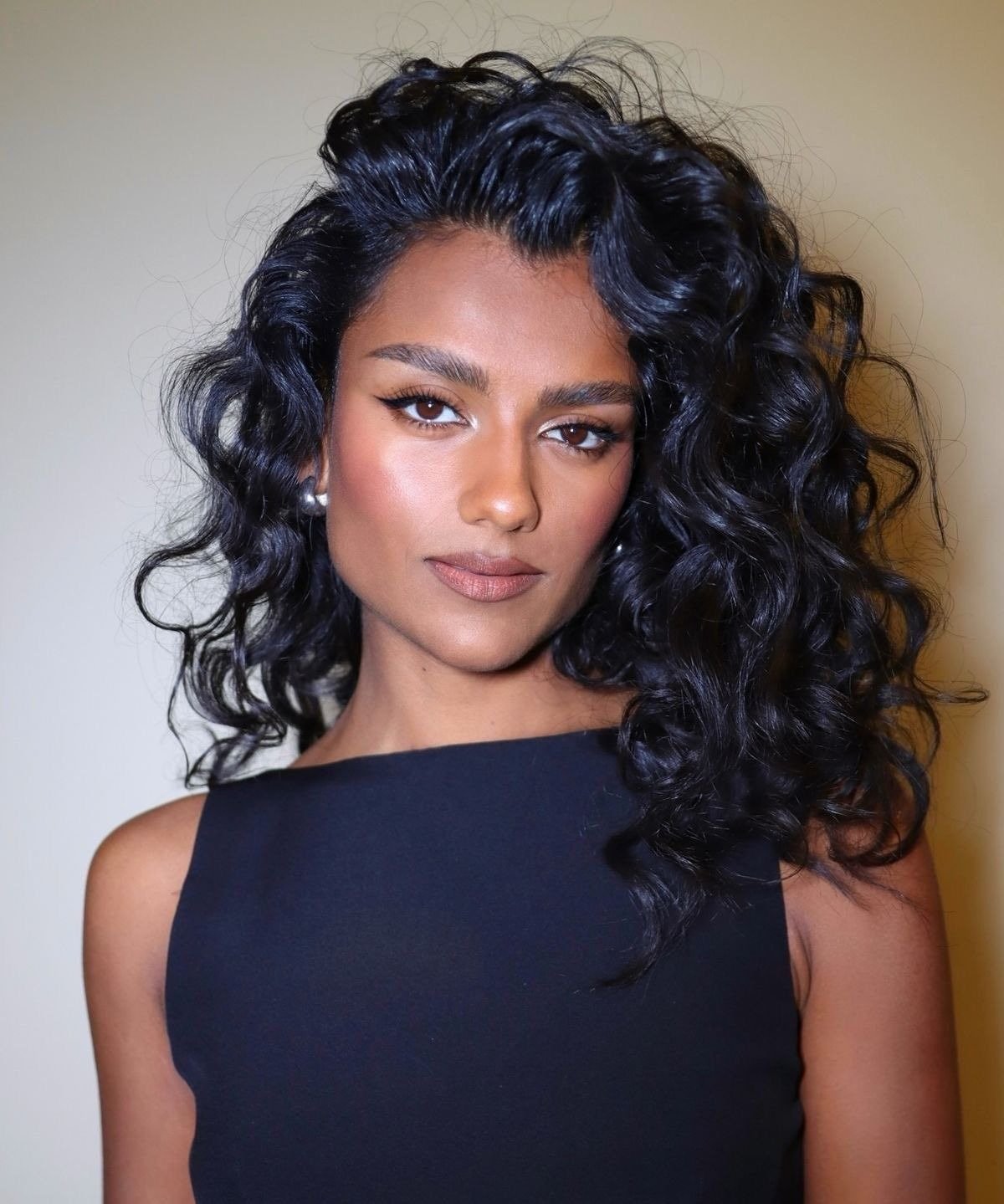 Modelo posa com cabelo ondulado volumoso e brilhoso, vestido preto minimalista e maquiagem natural realçando a beleza.