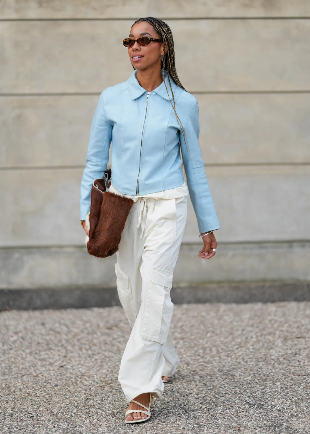 Modelo caminha confiante usando calça cargo branca de tecido leve com bolsos laterais, jaqueta azul clara e bolsa marrom.