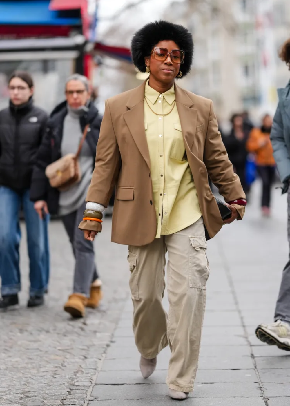 Modelo caminha confiante com calça cargo bege de tecido leve, camisa amarela e blazer marrom, em estilo urbano sofisticado.