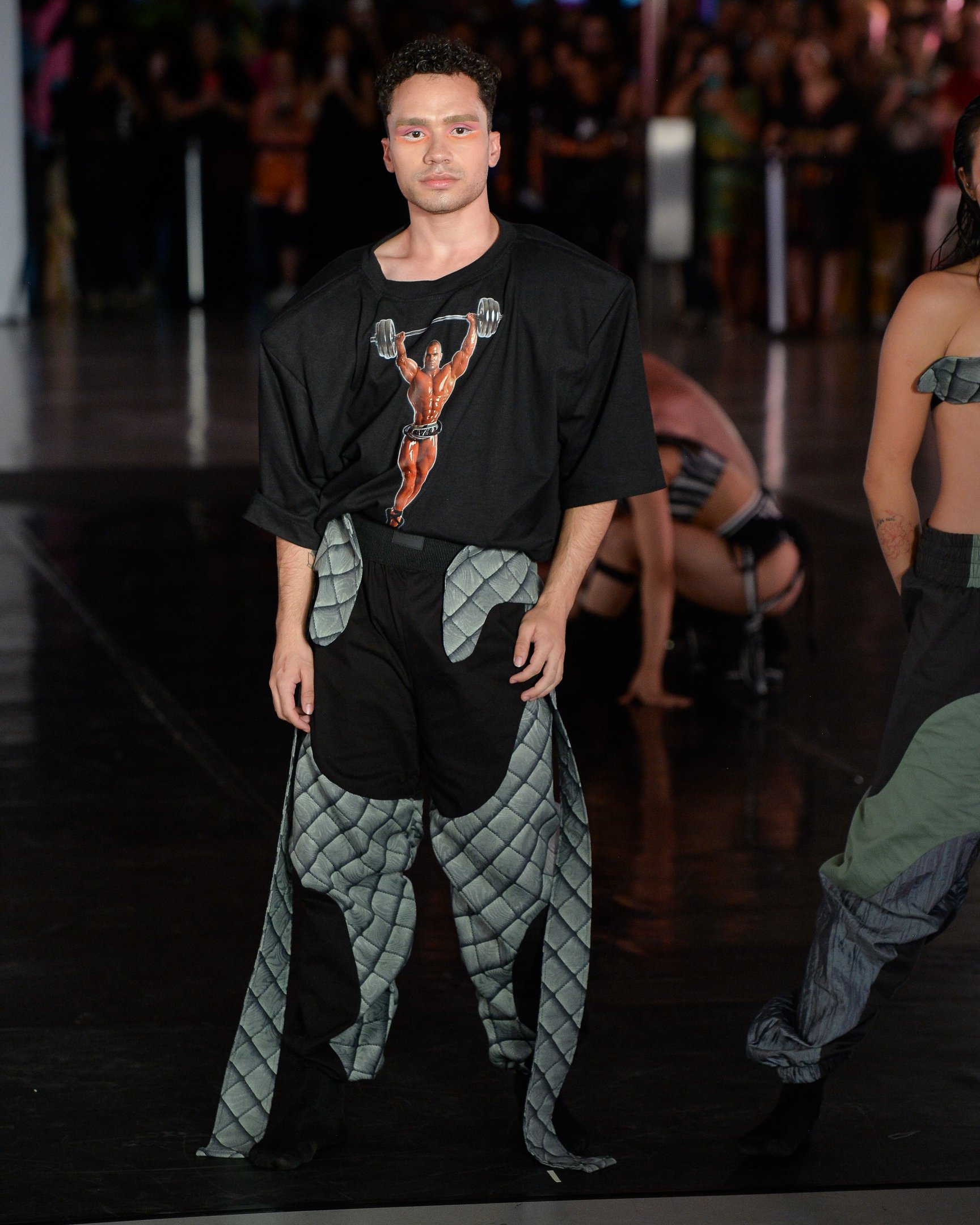 Modelo posa com camiseta preta oversized com estampa de fisiculturista e calça ampla com sobreposições em tecido matelassê