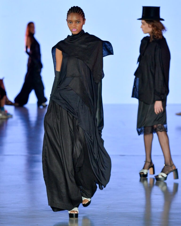 Modelo caminha com postura firme usando look preto fluido e assimétrico em tecidos leves, criando atmosfera dramática na
