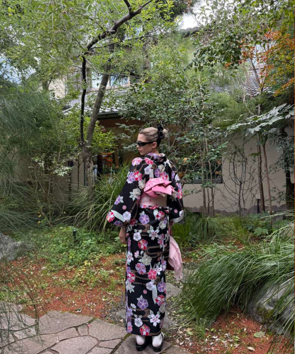 Mulher posa em jardim com kimono preto floral em tons de rosa e lilás, obi rosa e acessórios que reforçam dicas de Tóquio.