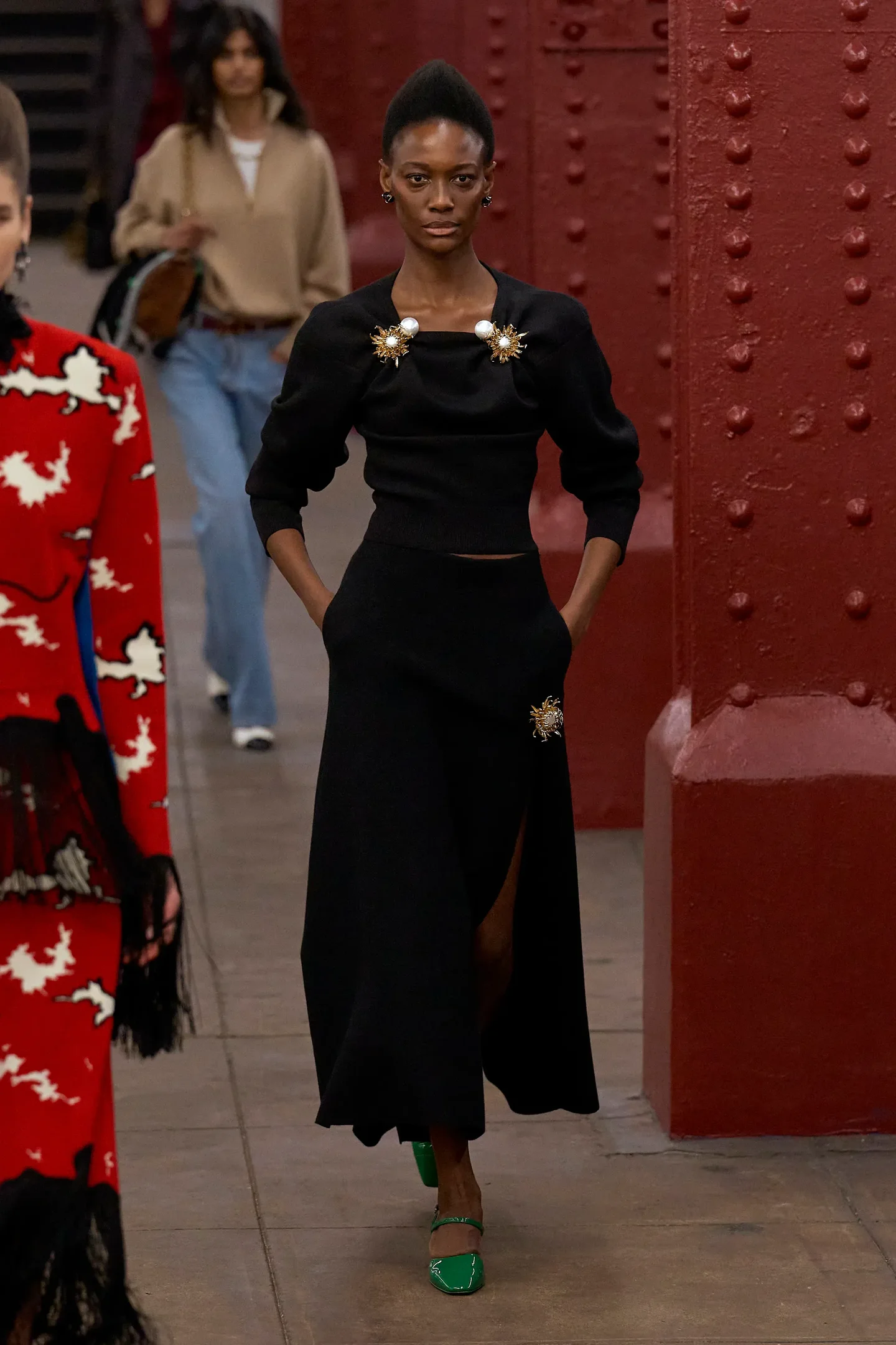 Modelo caminha com postura confiante na Chanel Métiers d’art 2026 usando conjunto preto de tricô com broches dourados.