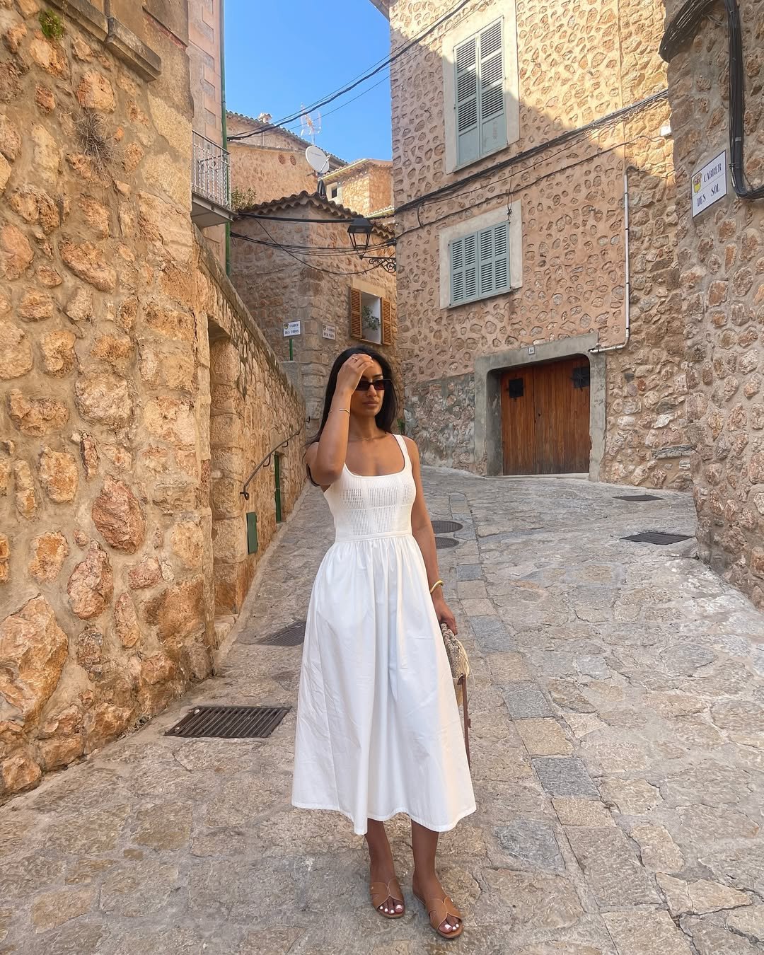 Modelo posa em vestido midi branco com corpete texturizado e saia fluida, combinando sandálias caramelo em cenário rústico