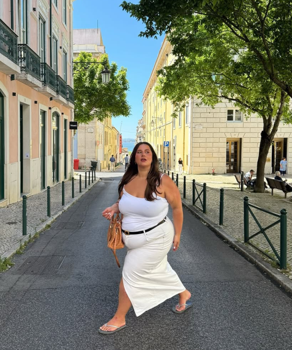 Modelo caminha confiante em rua ensolarada usando saia midi branca com fenda, regata branca e bolsa caramelo.