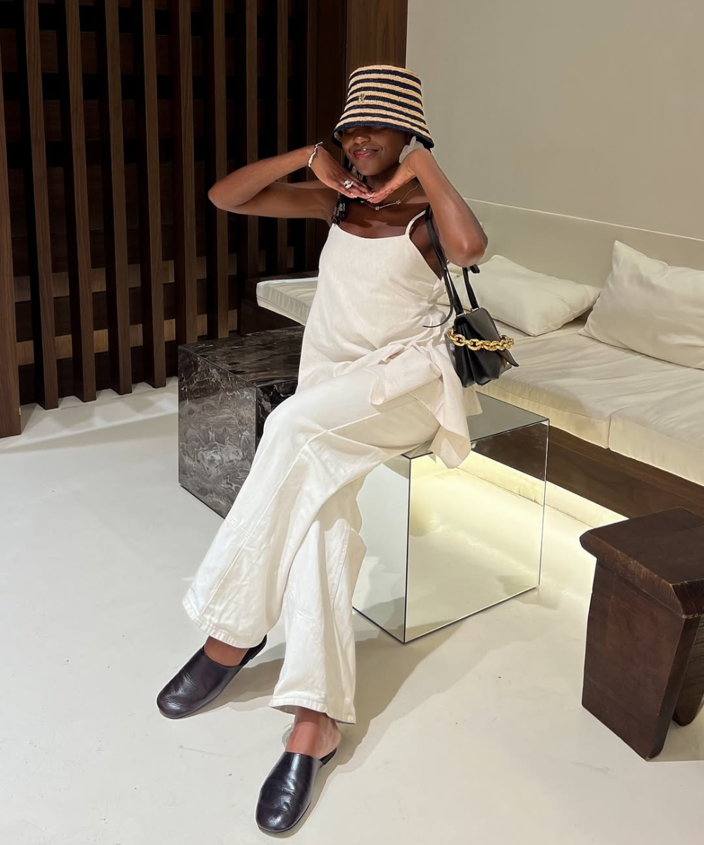 Modelo senta descontraída com look off-white fluido, chapéu listrado e bolsa preta, destacando a cor do ano da Pantone.