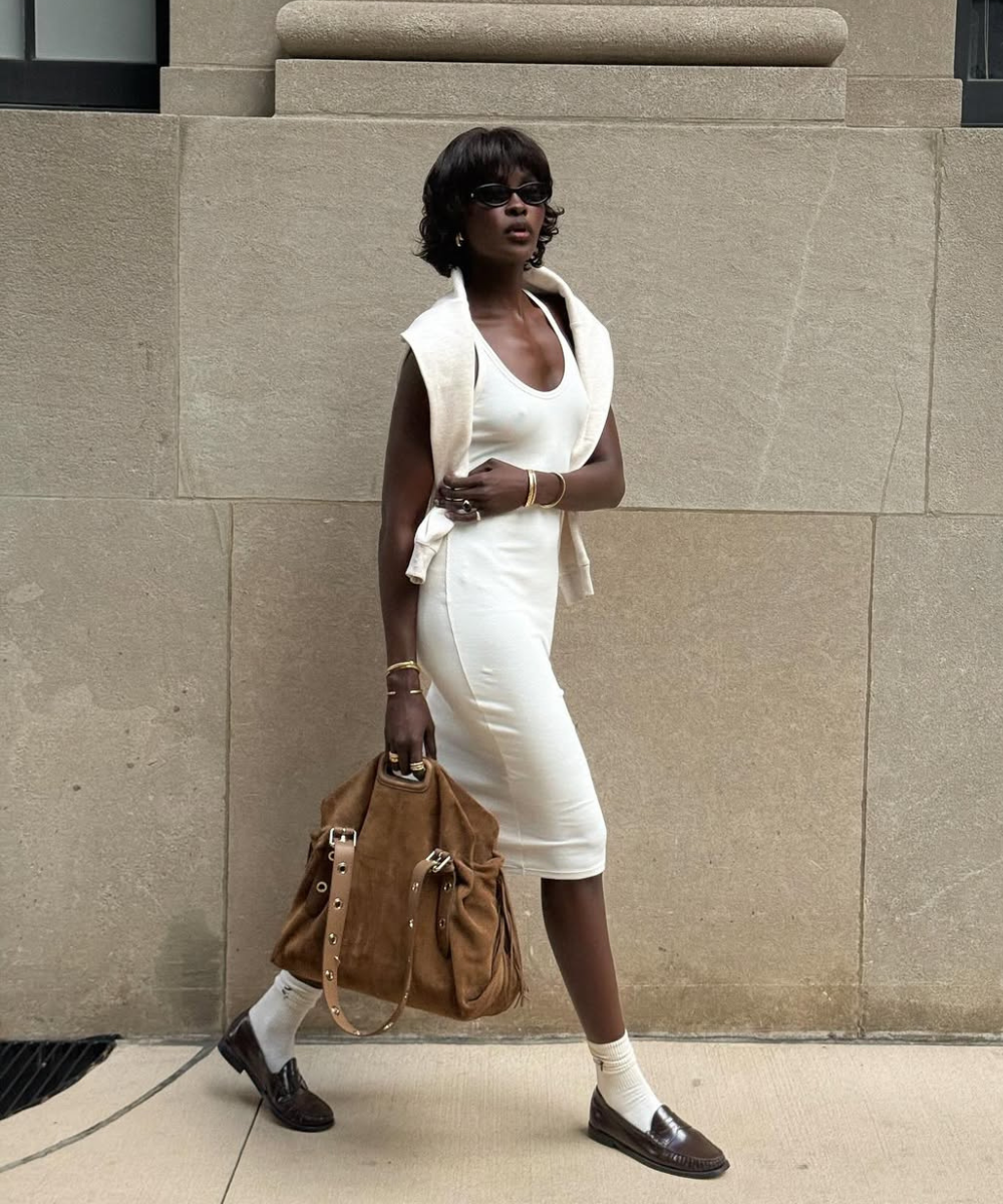 Modelo caminha com vestido branco justo em malha, suéter creme sobre os ombros e bolsa caramelo em camurça.
