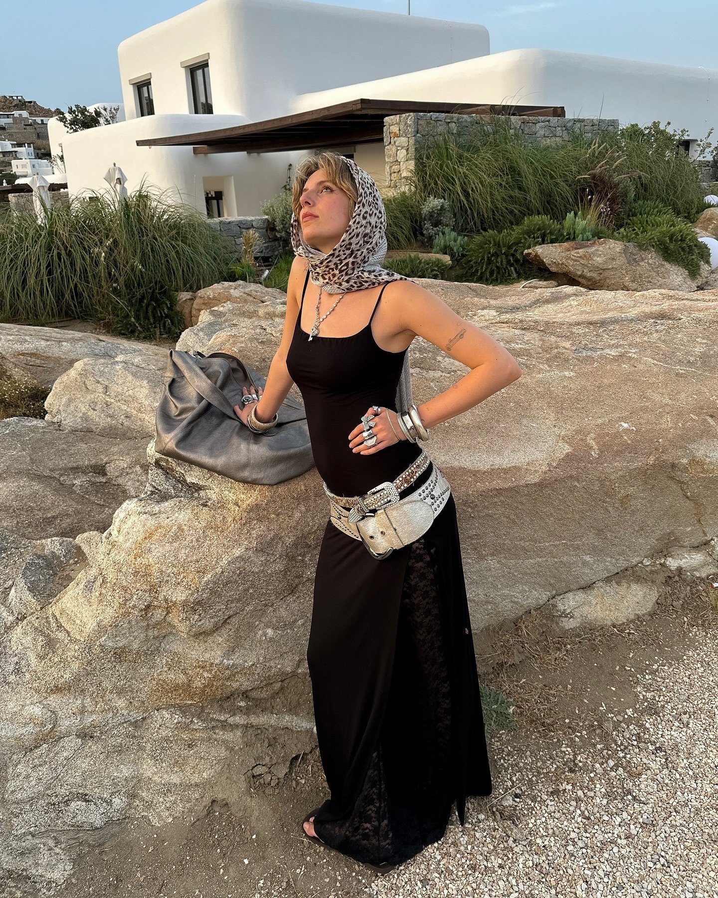 Modelo posa com vestido longo preto justo de alças finas e fenda lateral rendada, usando cinto largo prateado com detalhes