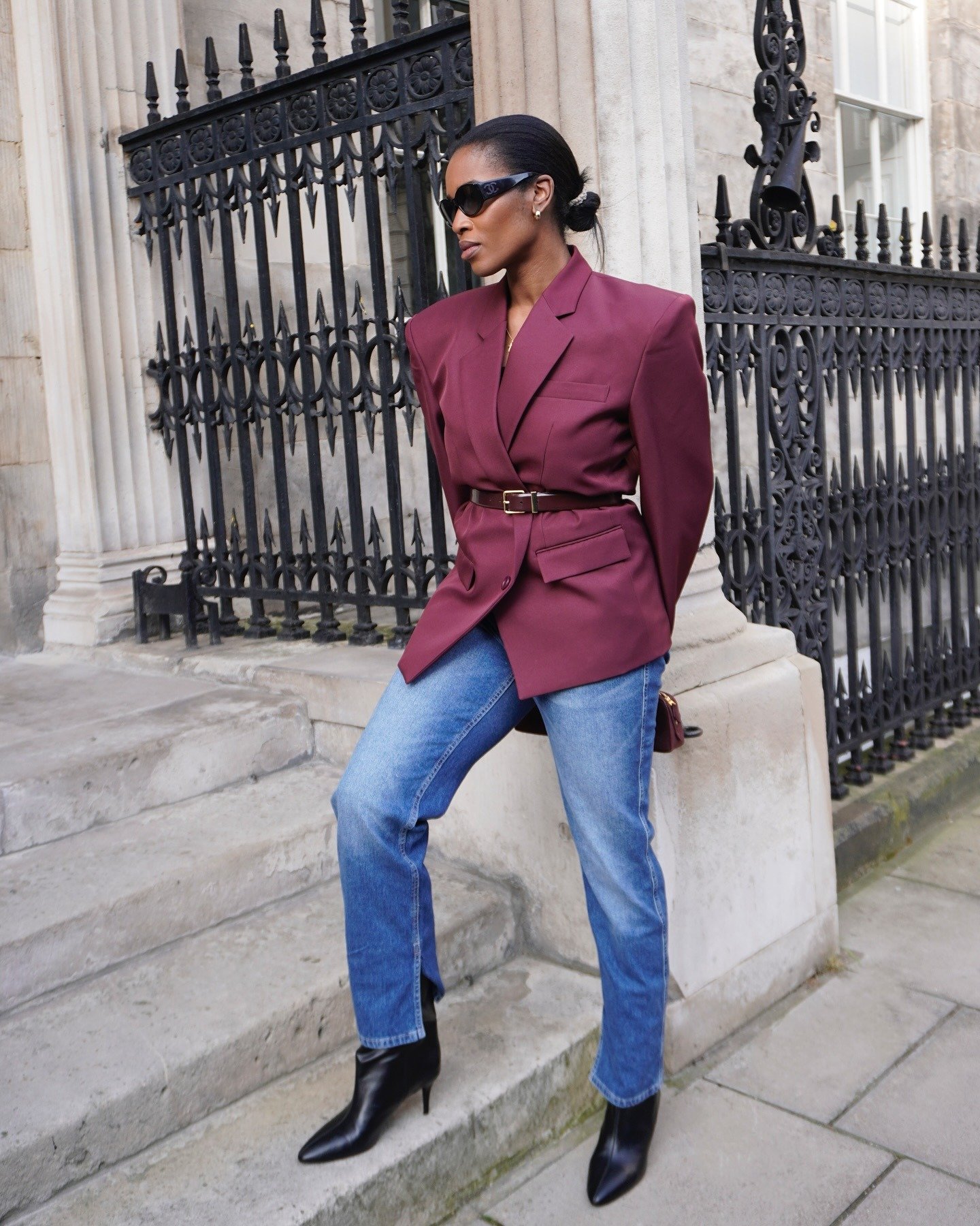 Modelo posa com blazer vinho oversized acinturado por cinto marrom fino, jeans azul e bota preta, revelando como usar cinto