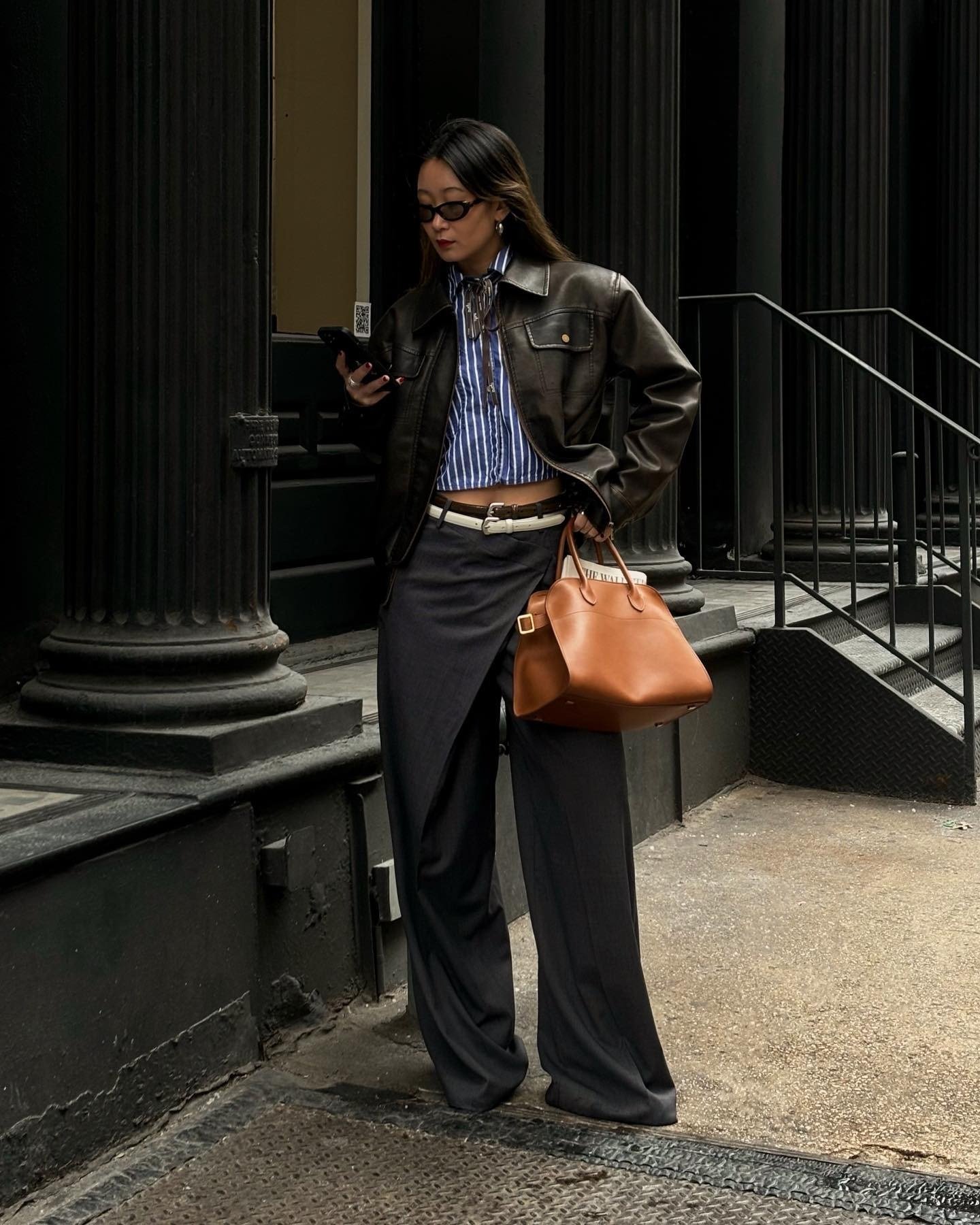 Modelo usa cinto branco sobre calça preta de alfaiataria com fenda, jaqueta de couro marrom e camisa listrada, em pose