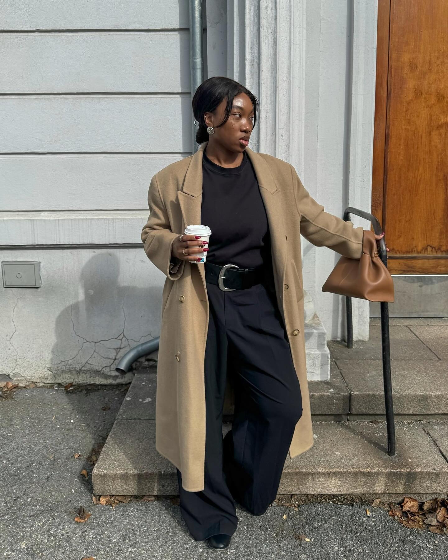 Modelo posa com casaco bege oversized, blusa preta, calça pantalona e cinto largo preto que define a cintura com elegância.