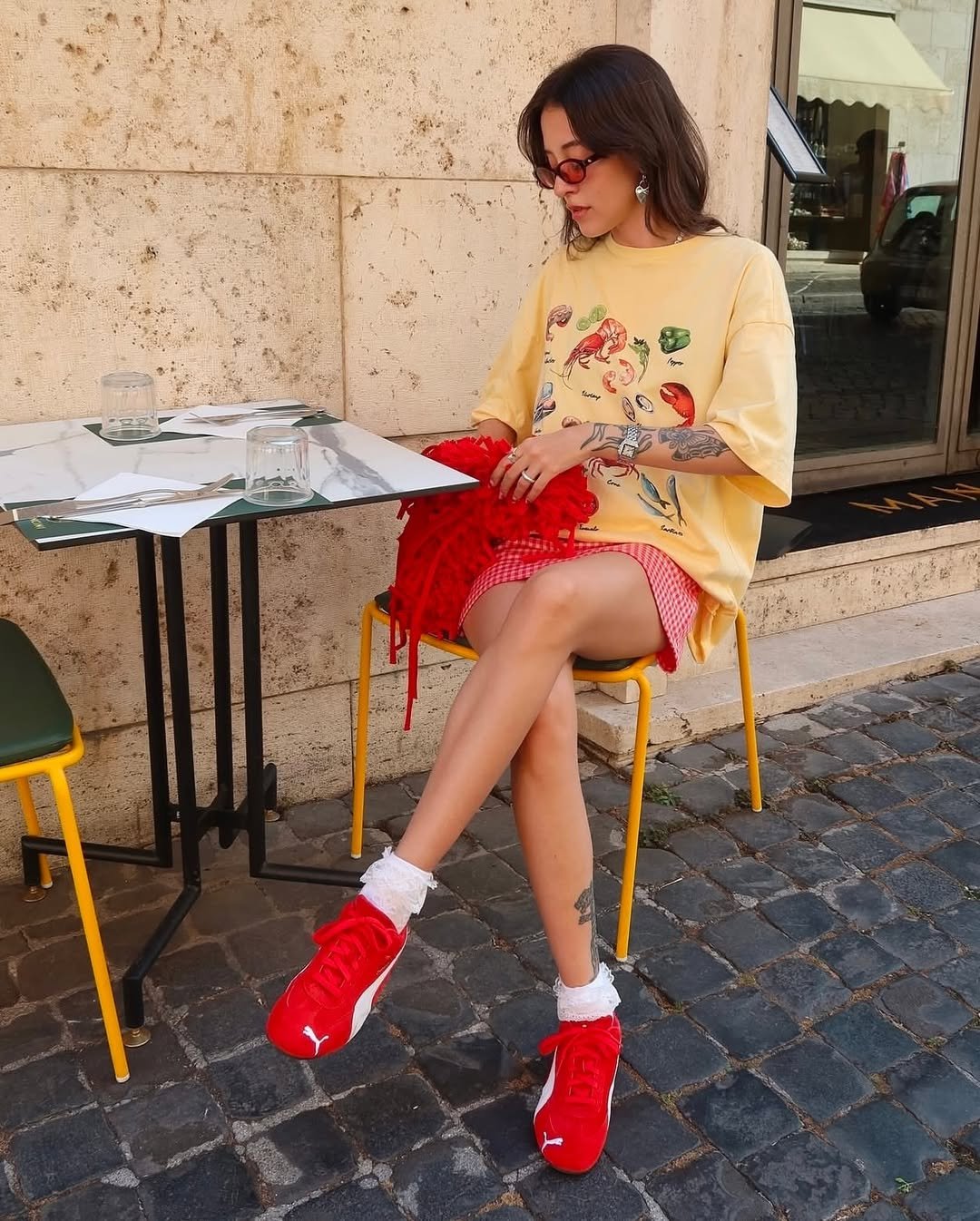 Mulher sentada em mesa externa usa camiseta amarela e tênis vermelhos, cores de Gêmeos para 2026, com estilo descontraído.
