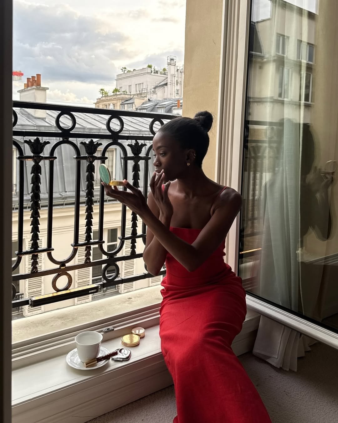 Modelo senta à janela em vestido vermelho vibrante de alças finas, refletindo elegância e sofisticação na cor do ano 2026.