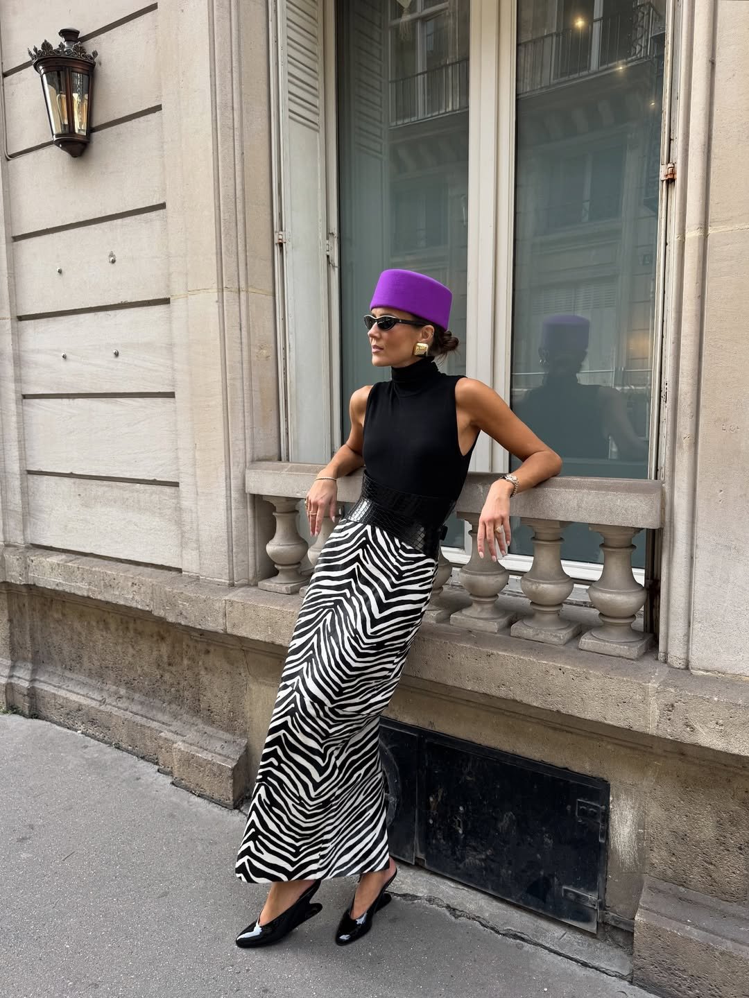 Mulher com saia zebrada e chapéu violeta, cor de Aquário para 2026, posa elegante encostada em prédio clássico.