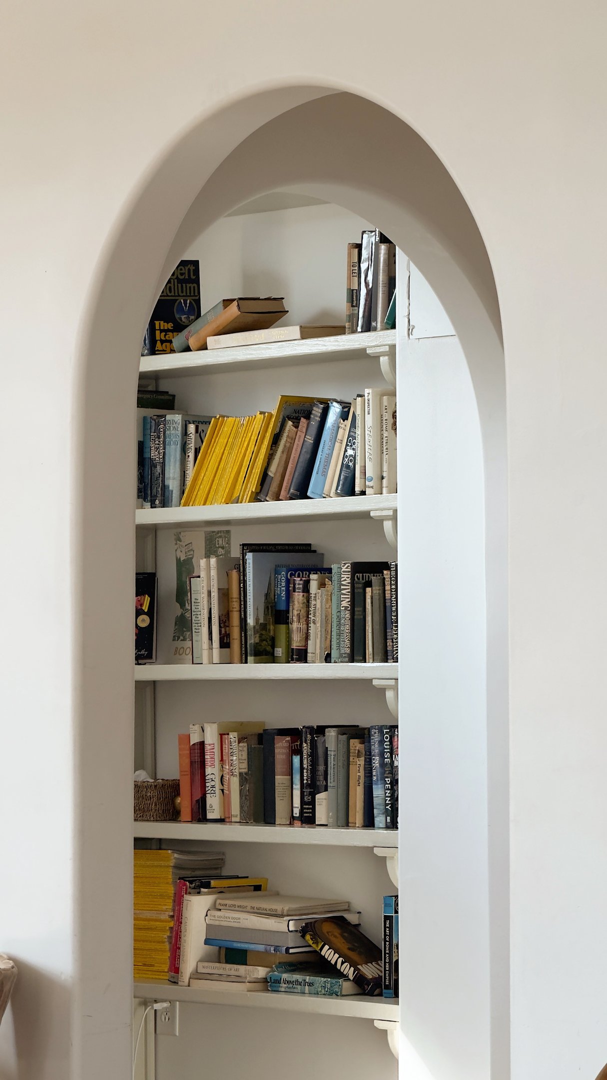 Estante branca com livros analógicos de capas variadas em tons neutros e amarelos, criando ambiente acolhedor e intelectual.