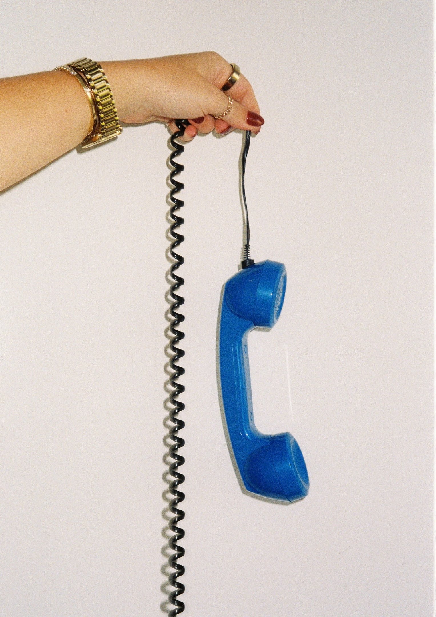 Mão feminina com acessórios dourados segura telefone analógico azul com fio espiral preto, em fundo branco minimalista.