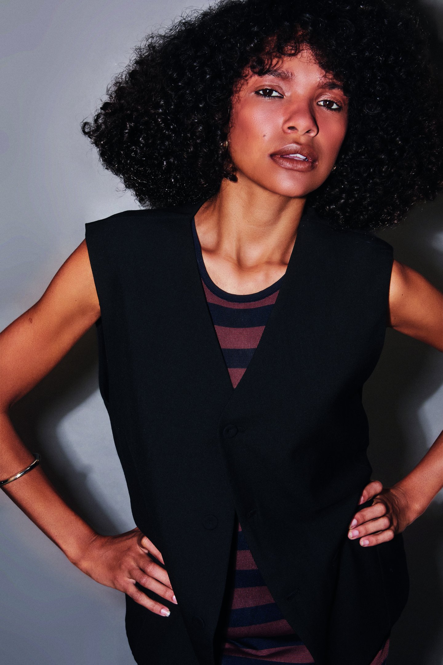 Modelo posa com cortes de cabelo da moda em fios cacheados volumosos, combinando colete preto estruturado e blusa listrada.