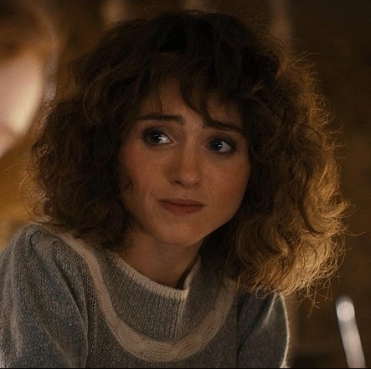 Corte de cabelo de Stranger Things com volume e cachos soltos em tom castanho, combinando com suéter cinza em ambiente
