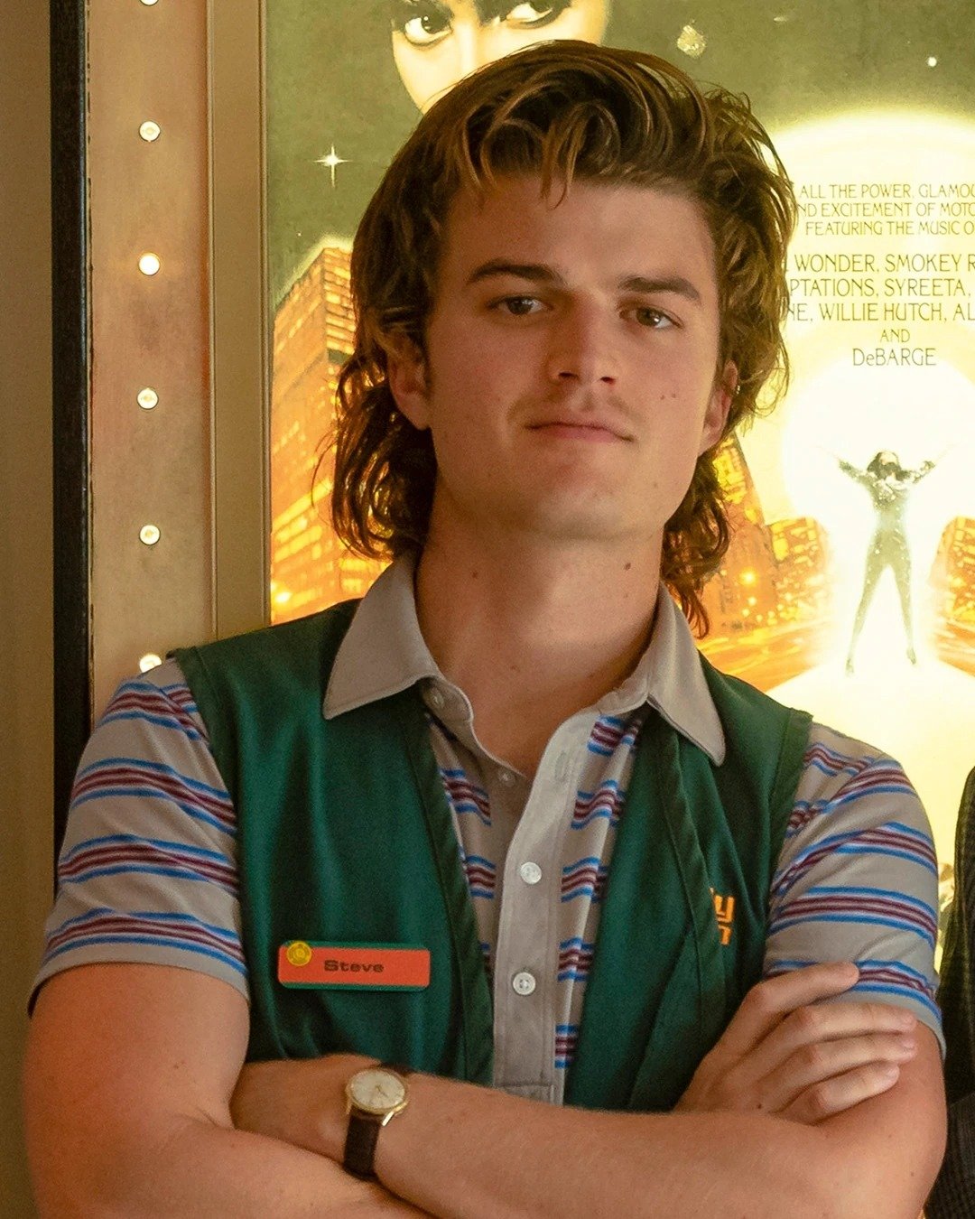 Corte de cabelo de Stranger Things com mullet ondulado, camisa polo listrada azul e cinza, colete verde e relógio clássico,