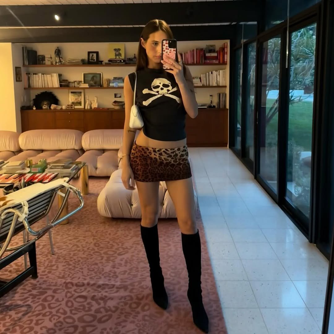 Devon Lee Carlson exibe estilo com regata preta justa de caveira, minissaia animal print e botas pretas altas, em pose