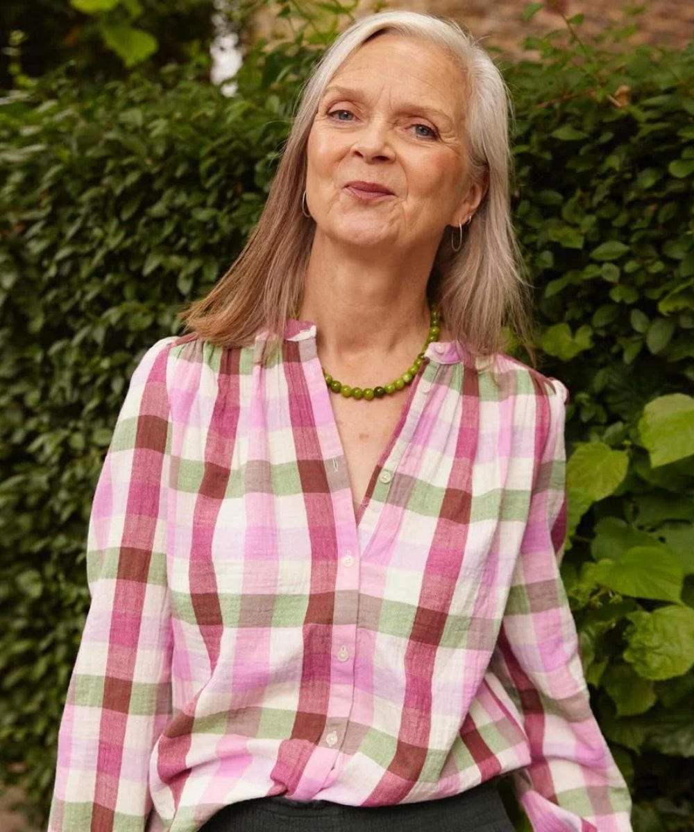 Mulher posa sorrindo com blusa xadrez rosa, verde e branca em tecido leve, destacando a melhor base para pele madura.