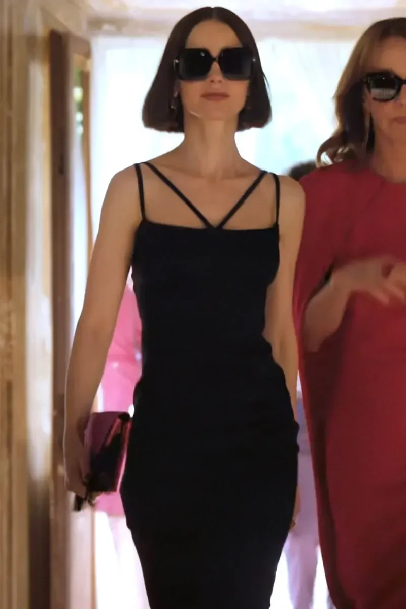 Emily em Paris exibe vestido preto justo com alças finas cruzadas, óculos oversized e clutch vinho, caminhando confiante