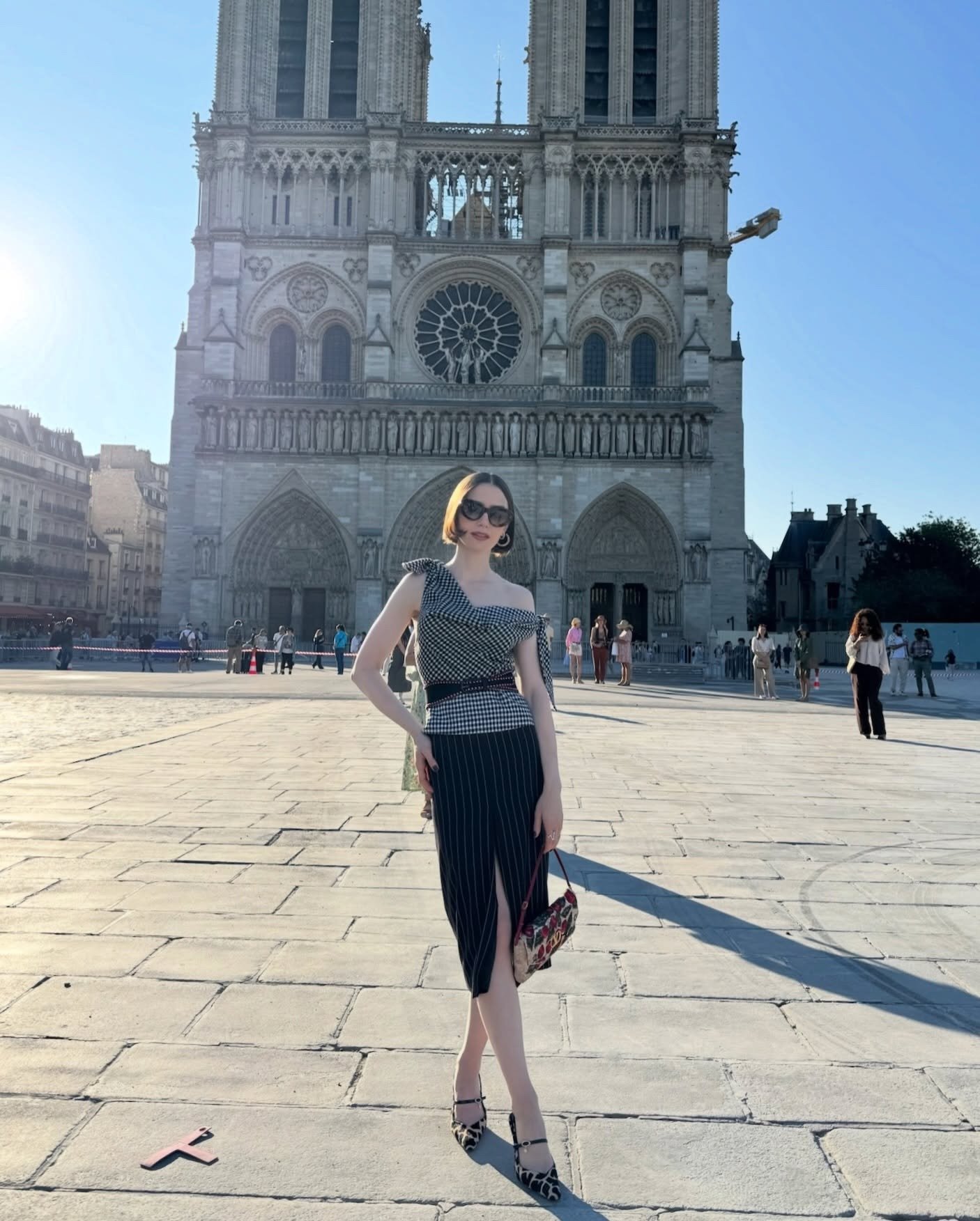 Mulher posa em frente à Notre-Dame com blusa xadrez assimétrica, saia lápis listrada, bolsa floral e scarpin animal print.
