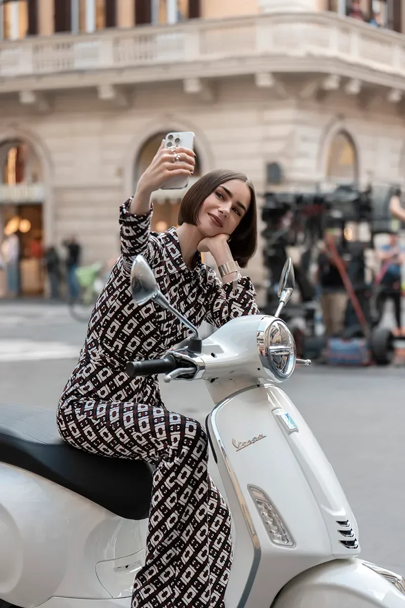 Modelo posa sentada em scooter branca com macacão estampado em preto e branco, cabelo curto e expressão confiante no estilo