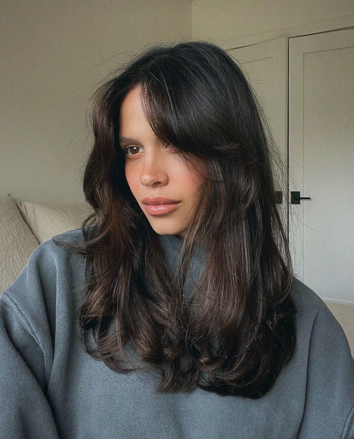 Modelo posa com corte de cabelo 2026 em camadas longas e franja desfiada, cabelos castanho escuro e look casual cinza