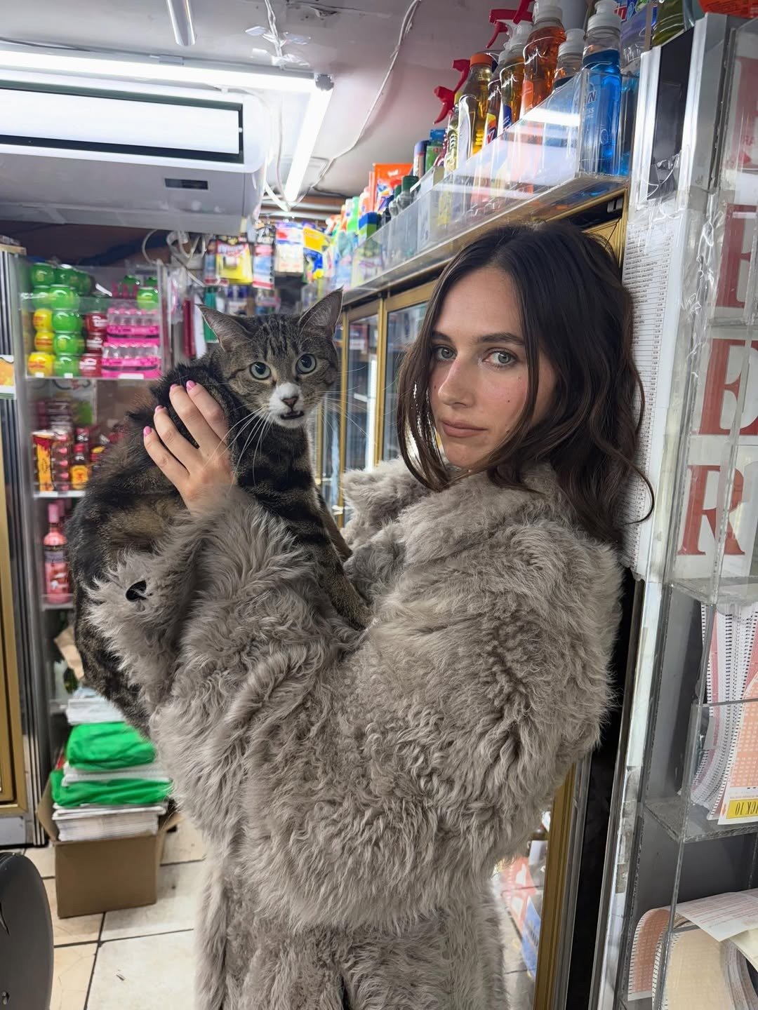 Devon Lee Carlson exibe estilo com casaco peludo oversized bege, cabelo solto e natural, posando segura um gato em clima