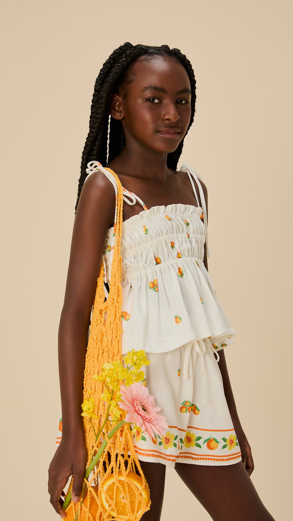 Modelo posa com blusa de alças finas e shorts em algodão branco estampados com frutas e flores, bolsa de crochê laranja e