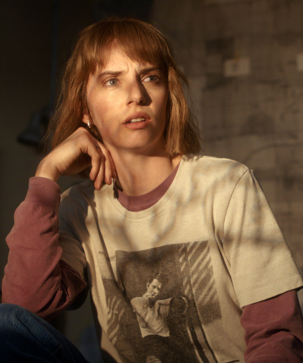 Mulher com camiseta Stranger Things off-white sobre manga longa vinho, posando pensativa em luz quente e ambiente neutro.