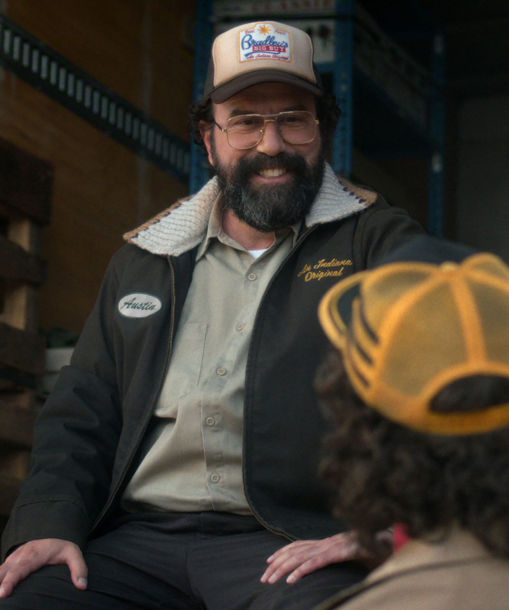 Personagem de Stranger Things sorri usando jaqueta preta com gola de lã, camisa bege e boné trucker bege e marrom.
