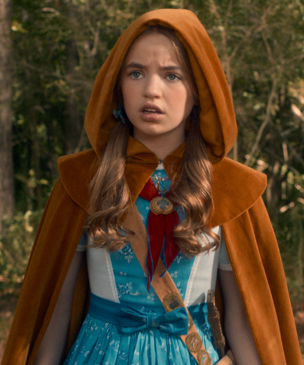 Personagem de Stranger Things usa capa marrom de veludo com capuz, vestido azul floral e faixa de escoteira.