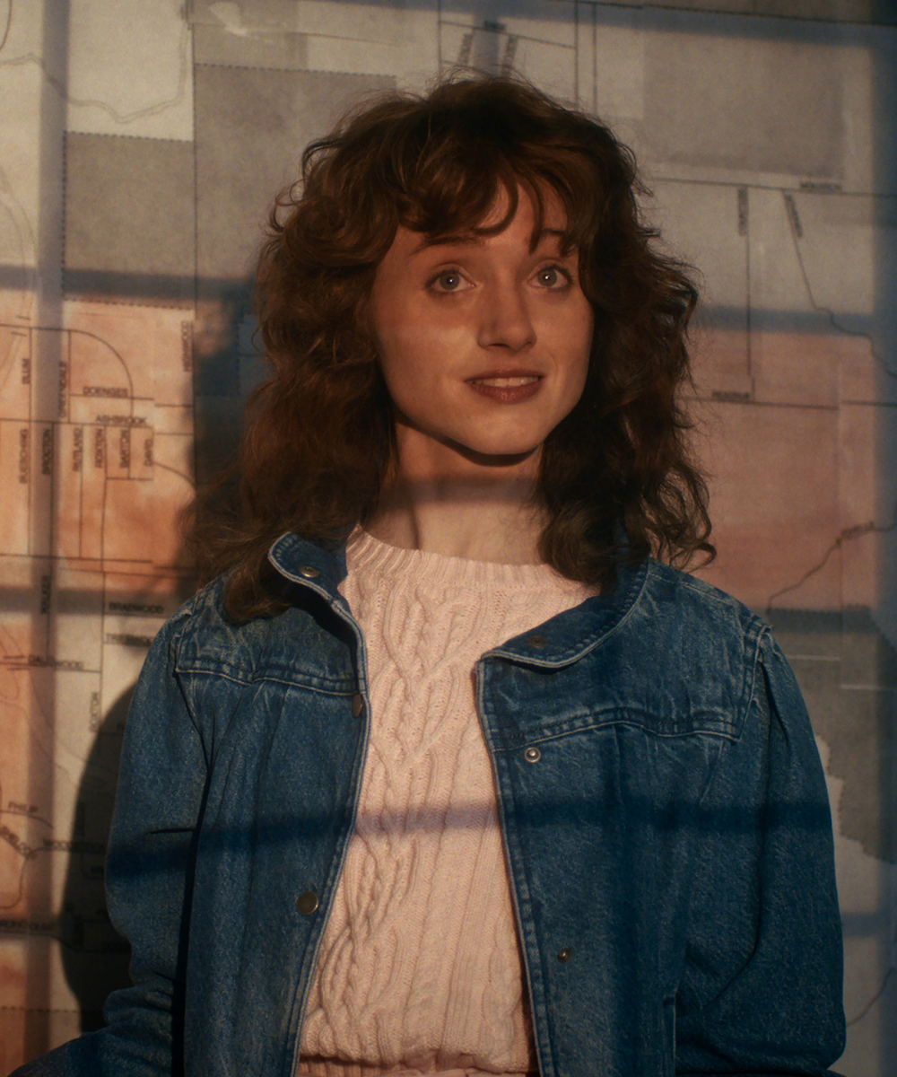 Mulher com jaqueta jeans oversized e suéter rosa de tricô posa sorrindo sob luz quente, estilo vintage de Stranger Things.
