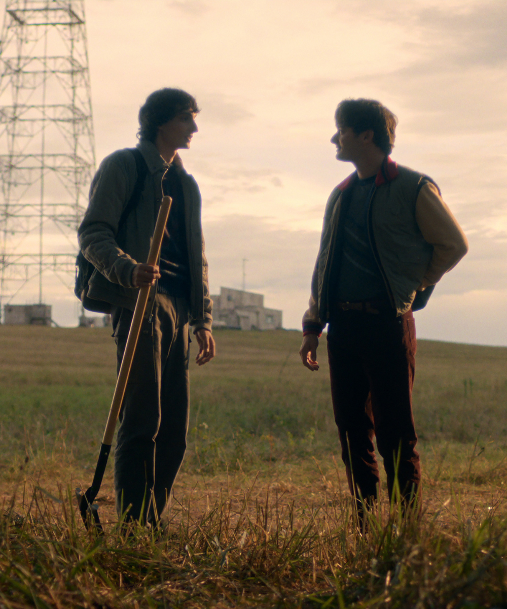 Dois jovens em campo aberto ao entardecer, um com jaqueta cinza clara e mochila segurando enxada, outro com jaqueta bomber.
