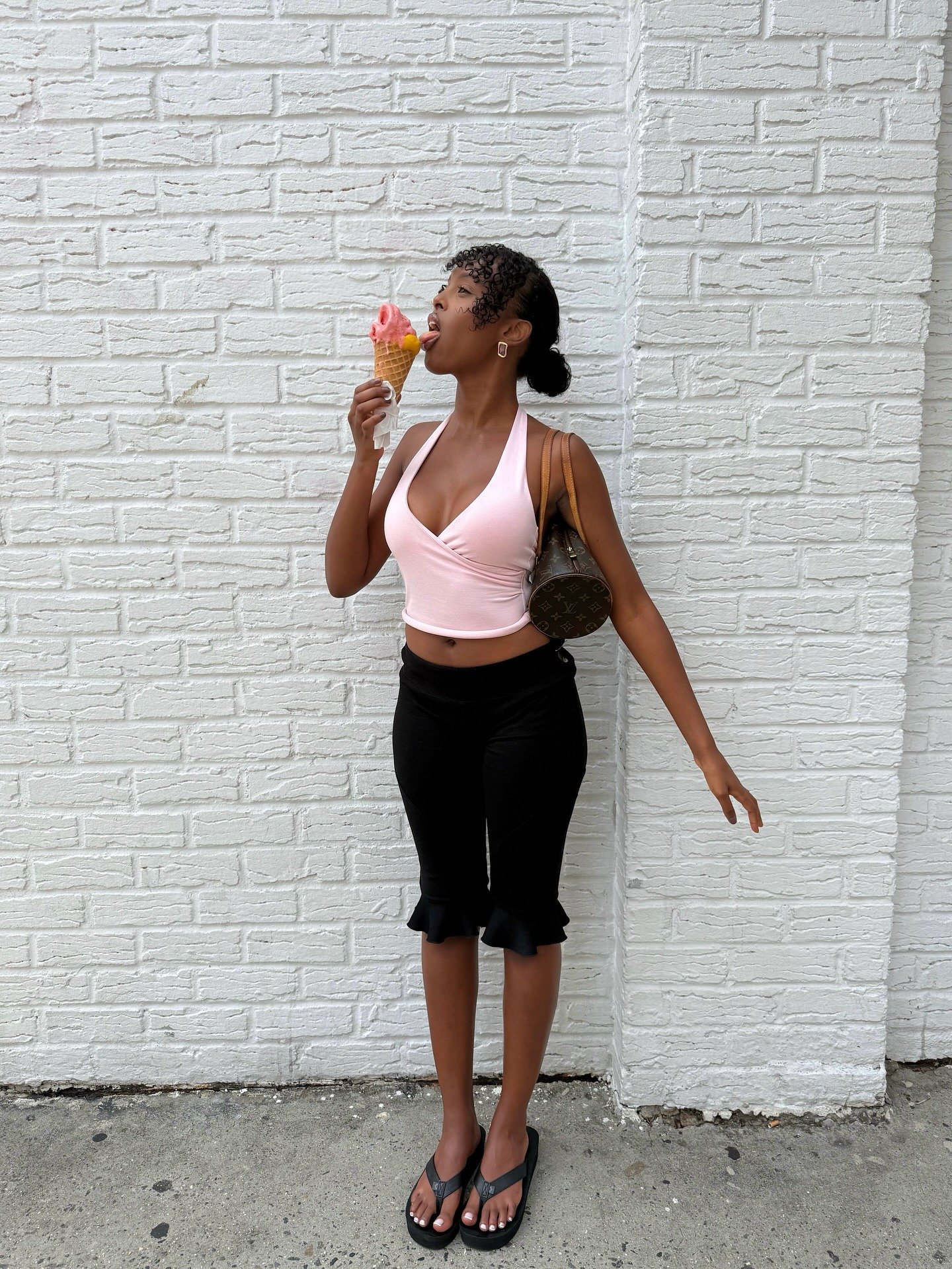 Modelo posa de forma descontraída com blusa rosa de alça, bermuda preta com babados e flatform preta, segurando sorvete rosa.