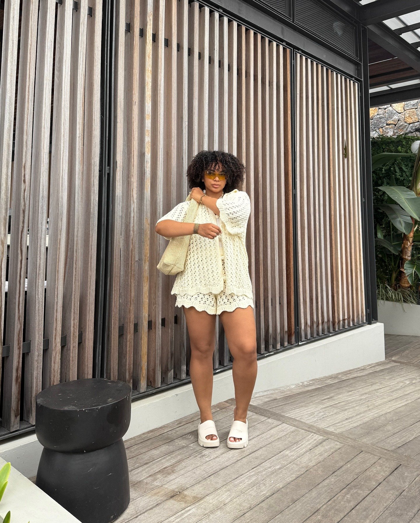 Modelo posa com look em crochê off-white composto por shorts e camisa de manga bufante, bolsa de palha e flatform branca, em