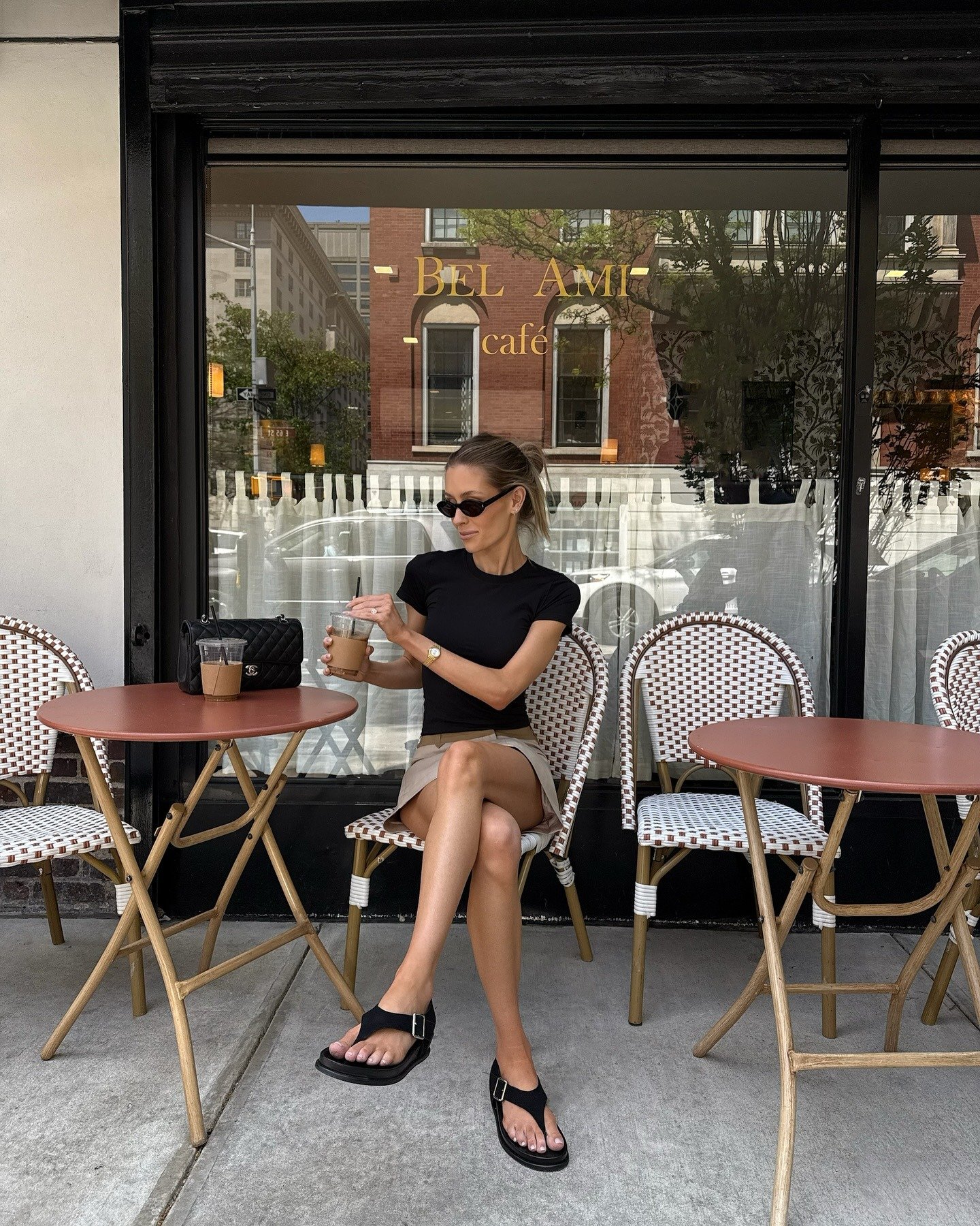 Modelo senta com postura relaxada em café urbano usando flatform preta de tiras largas, camiseta preta justa e saia bege,