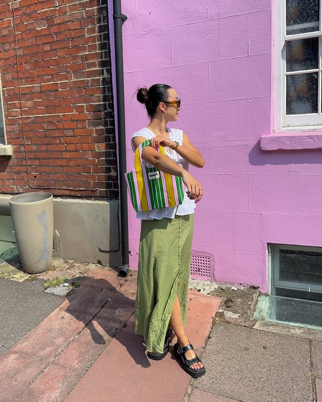 Mulher posa ao sol com saia verde longa, blusa branca e flatform preta de tiras, segurando bolsa listrada amarela e verde.