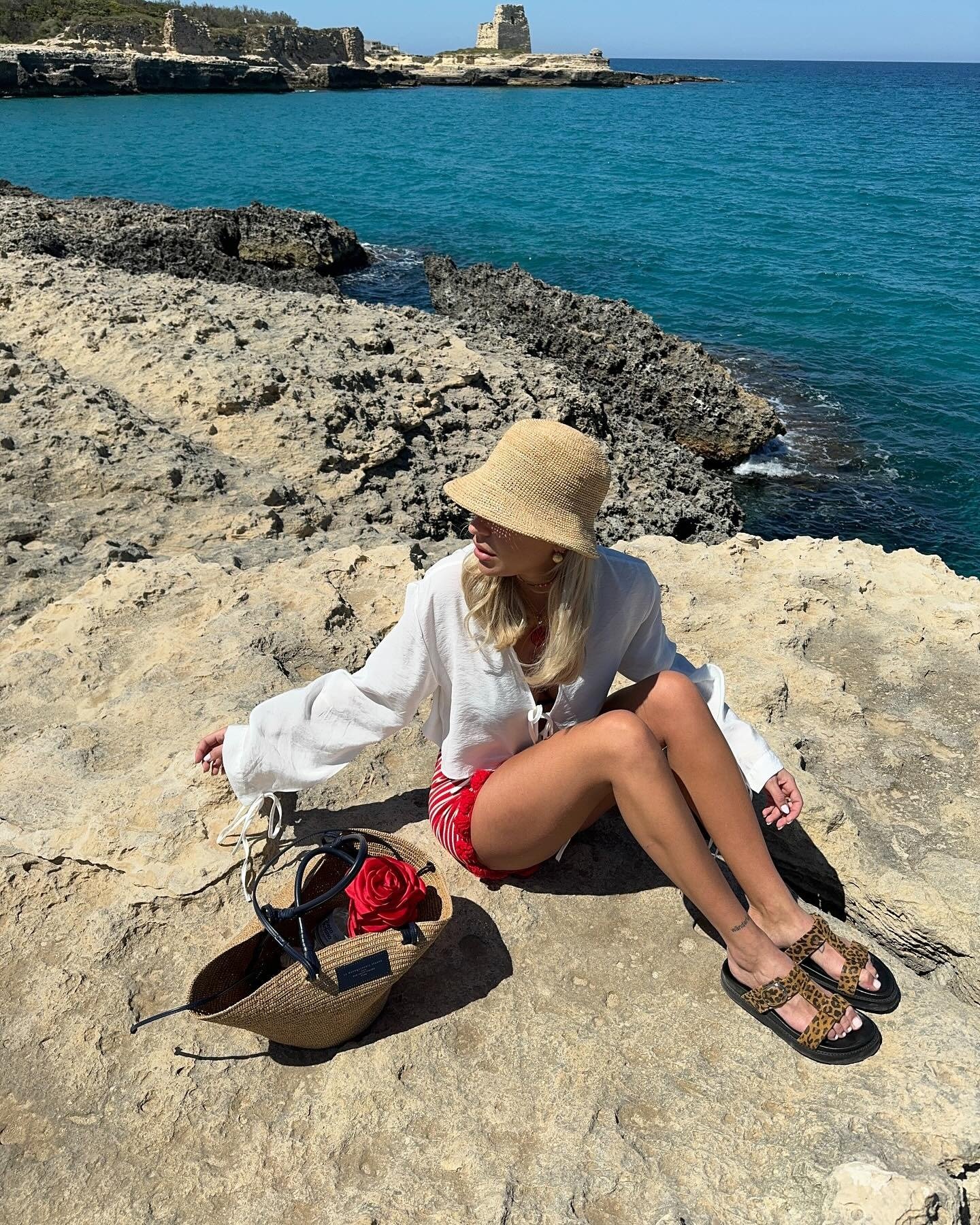 Mulher senta à beira do mar com flatform de tiras animal print, shorts vermelhos, blusa branca fluida e chapéu palha, clima