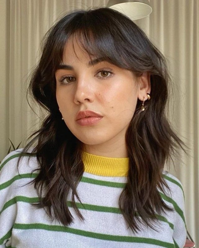 Corte de cabelo 2026 com camadas médias e franja desfiada em tom castanho natural, modelo posa com suéter listrado verde.