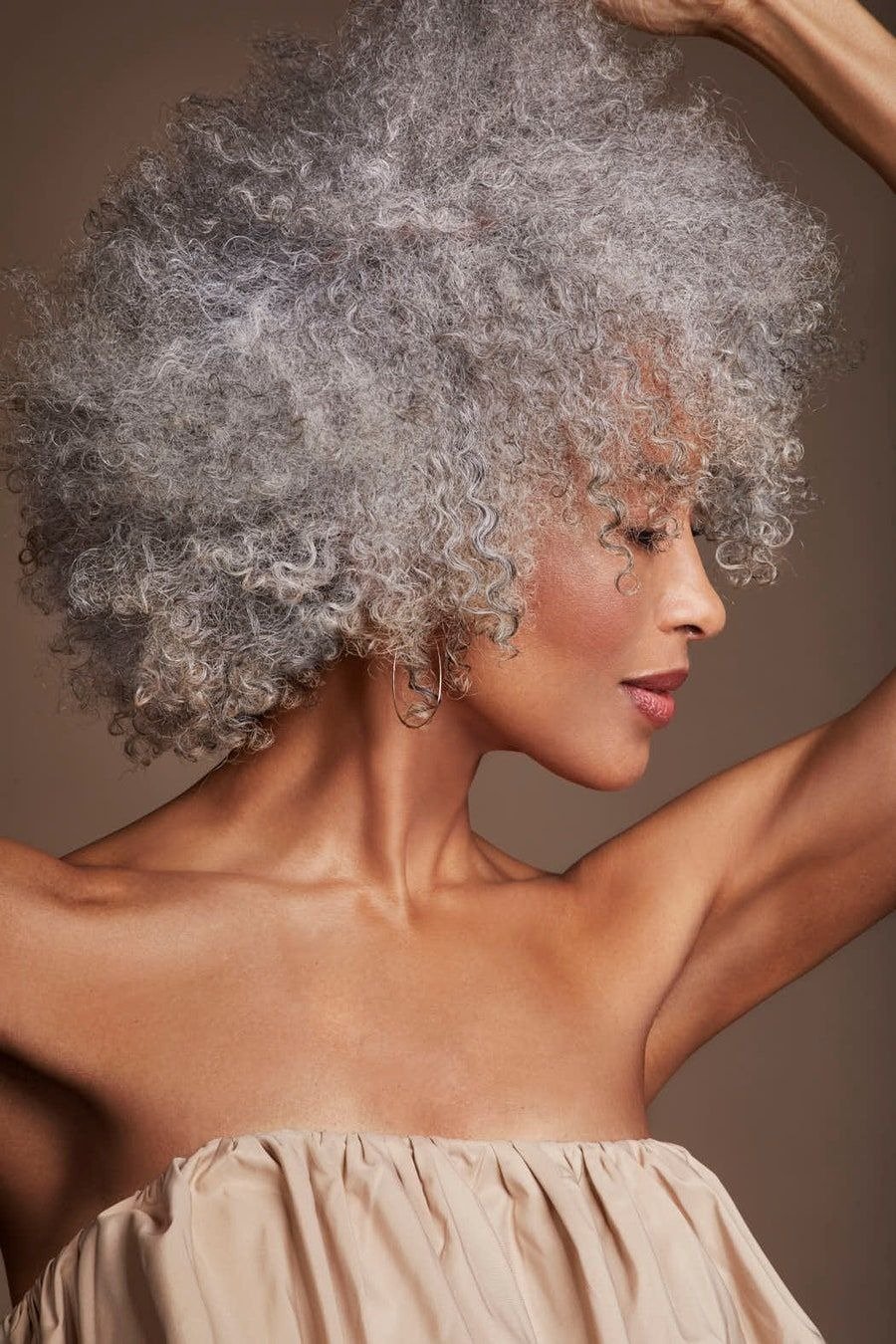 Mulher com grisalho cacheado volumoso, pele bronzeada, vestido tomara que caia bege e expressão serena em fundo neutro.