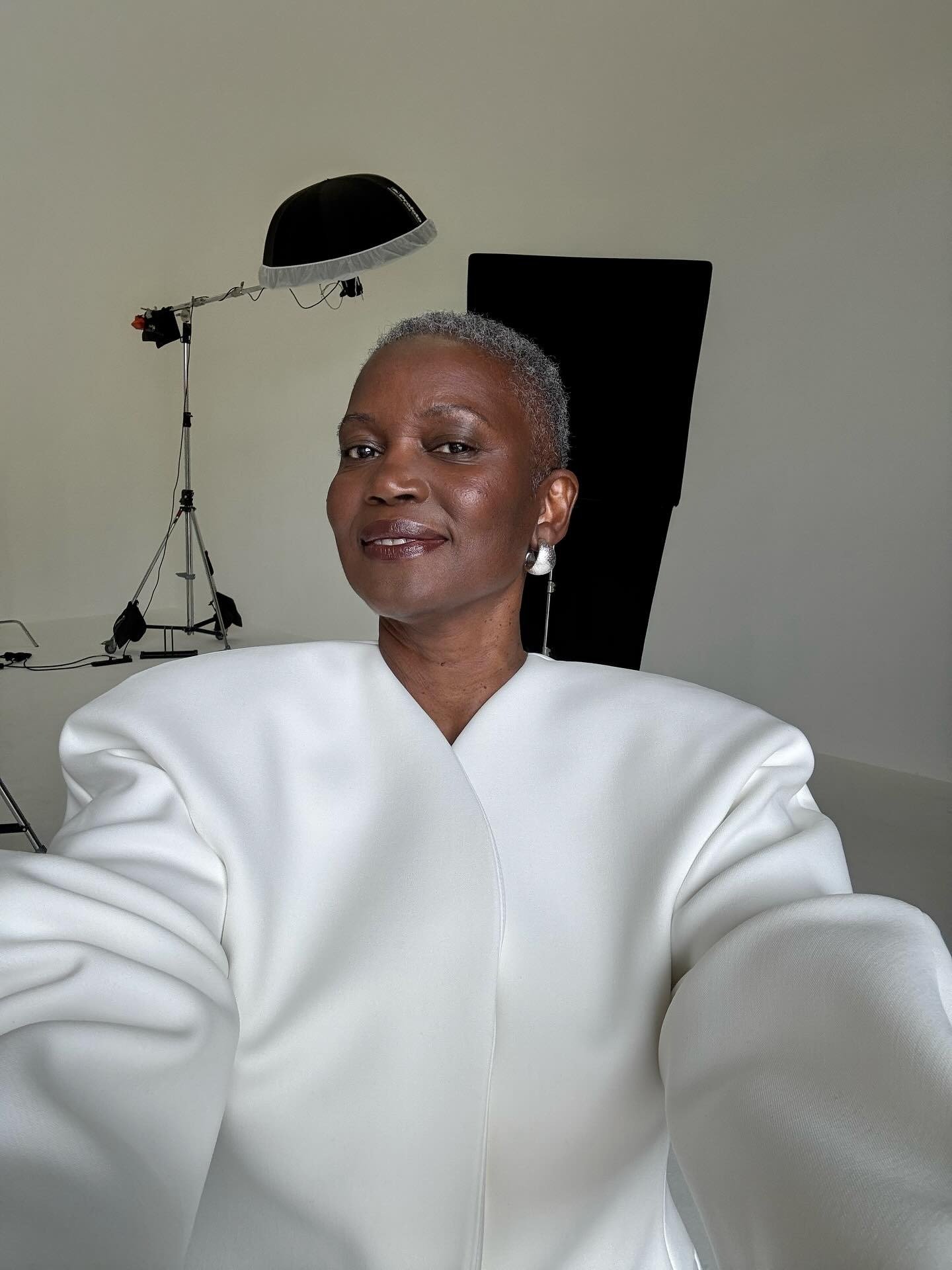 Grisalho cacheado exibe blazer branco oversized com ombros estruturados em estúdio minimalista, posando confiante e serena.