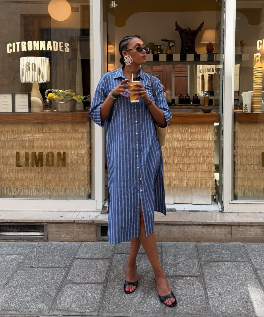 Modelo posa com vestido chemise midi listrado azul e branco, sandálias pretas e acessórios marcantes, em clima casual urbano.